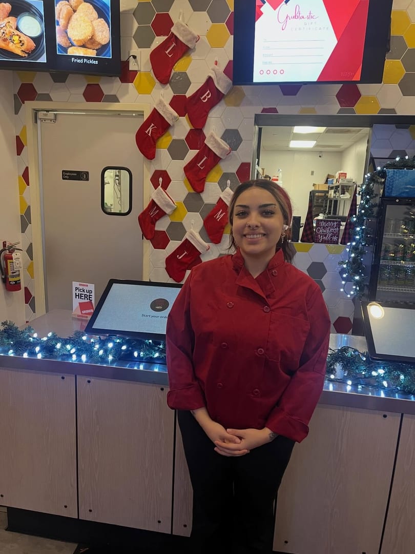 A smiling Grubtastic employee in a red chef coat stands at a counter decorated with Christmas lights and stockings.