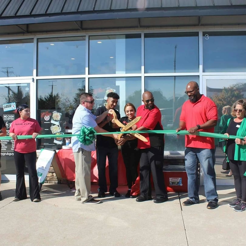 People in red shirts and a man in a blue shirt cutting a green ribbon for a grand opening.