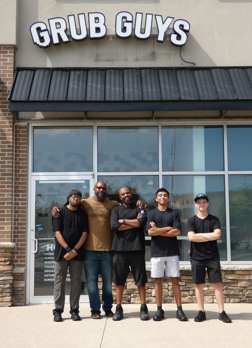 Five men, some in chef's attire, stand smiling in front of the Grub Guys restaurant.