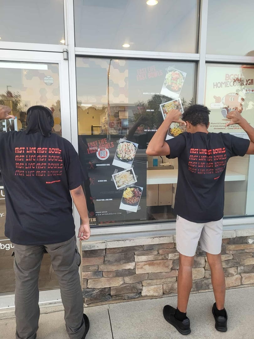 Two people in black "Fast Life Fast Bowls" shirts pose in front of a restaurant window.