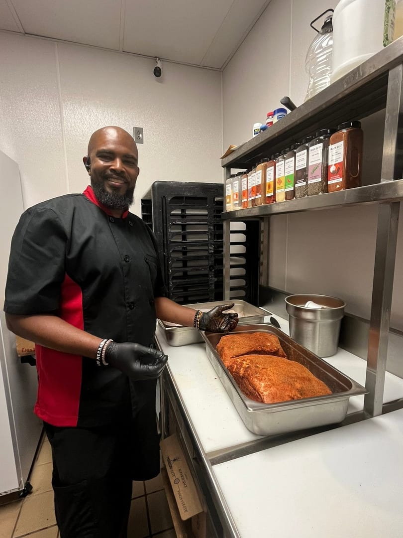 A chef in a black and red uniform stands next to two seasoned briskets in a commercial kitchen.