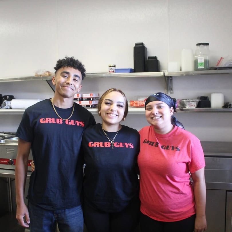 Three smiling Grub Guys employees, two women and one man, stand in a commercial kitchen.