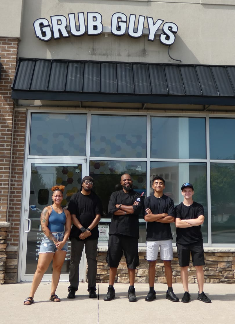 Five Grub Guys restaurant staff members stand smiling outside the storefront on a sunny day.