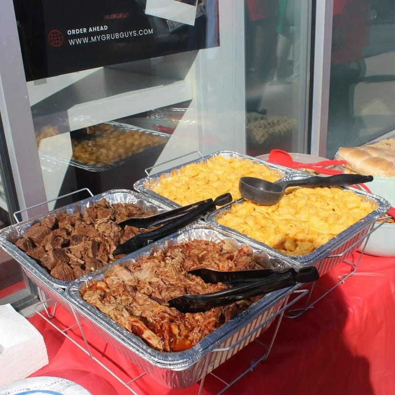 Catering spread with trays of mac and cheese, pulled pork, and beef, with a "My Grub Guys" sign.
