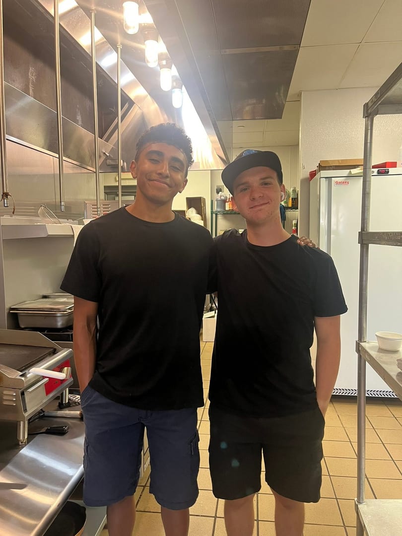 Two young men in black shirts and shorts smiling in a commercial kitchen.
