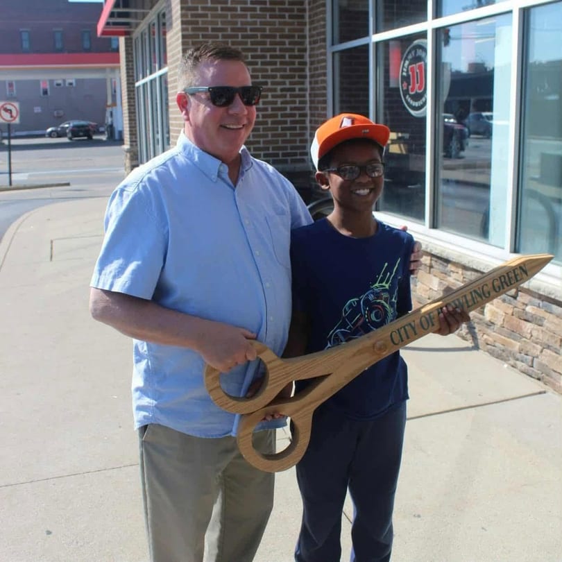 A man and a boy smiling, holding giant wooden scissors for a "City of Bowling Green" ribbon cutting.